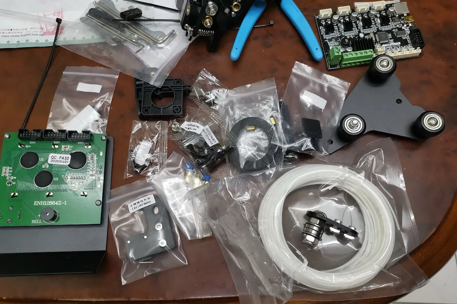 Parts of a Creality Ender 3 kit laid out in plastic bags on a wooden table: the mainboard, the LCD panel, the print head, a carriage plate with three wheels and a coil of white PTFE Bowden tube