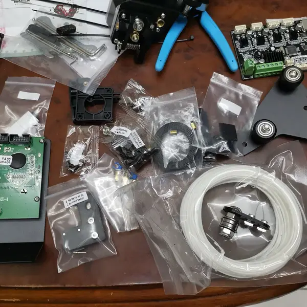Parts of a Creality Ender 3 kit laid out in plastic bags on a wooden table: the mainboard, the LCD panel, the print head, a carriage plate with three wheels and a coil of white PTFE Bowden tube