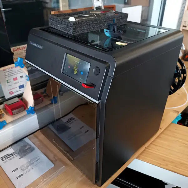 A black, fully enclosed Flashforge Adventurer 5M Pro on a wooden workbench, with its touchscreen lit, a blue Benchy and a foam accessory tray on its glass lid, and the quick start guide lying in front of it