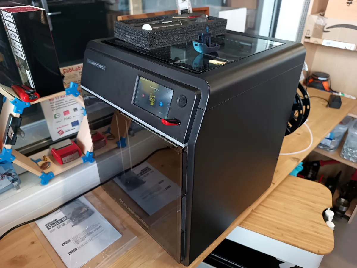 A black, fully enclosed Flashforge Adventurer 5M Pro on a wooden workbench, with its touchscreen lit, a blue Benchy and a foam accessory tray on its glass lid, and the quick start guide lying in front of it