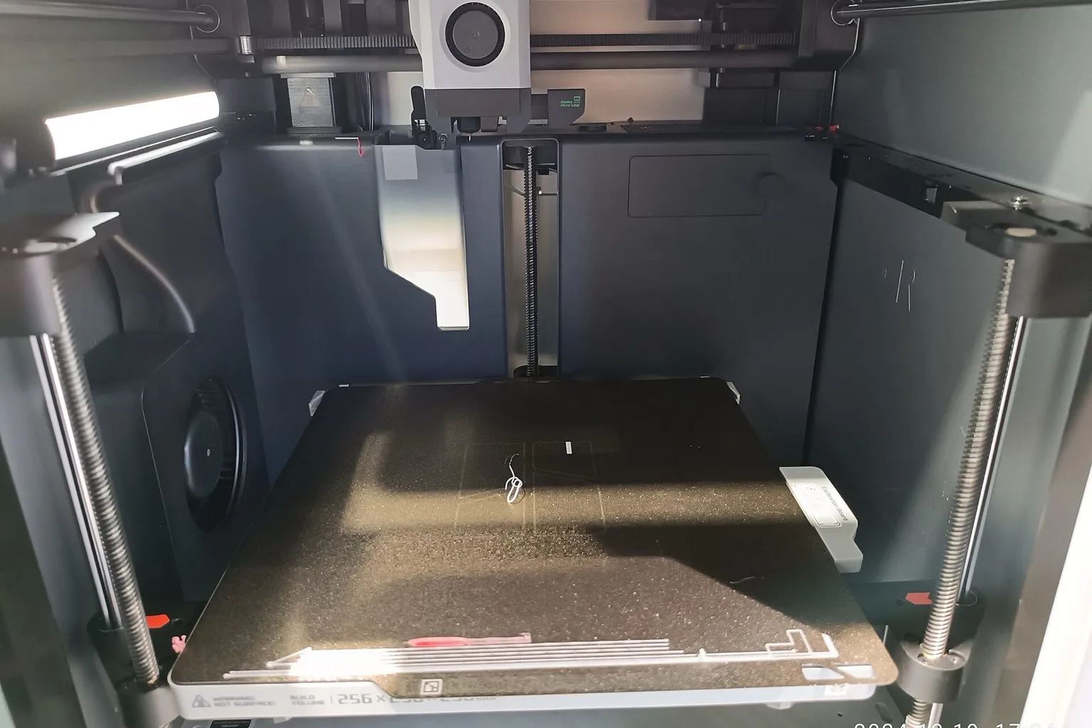 Inside an enclosed Bambu Lab printer on a showroom floor: a single-nozzle toolhead on the gantry above an empty textured build plate with a build-volume label on its front edge