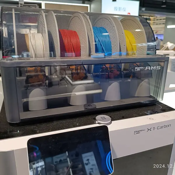 A Bambu Lab AMS holding white, red, blue and yellow filament spools on top of an X1-Carbon printer in a store showroom
