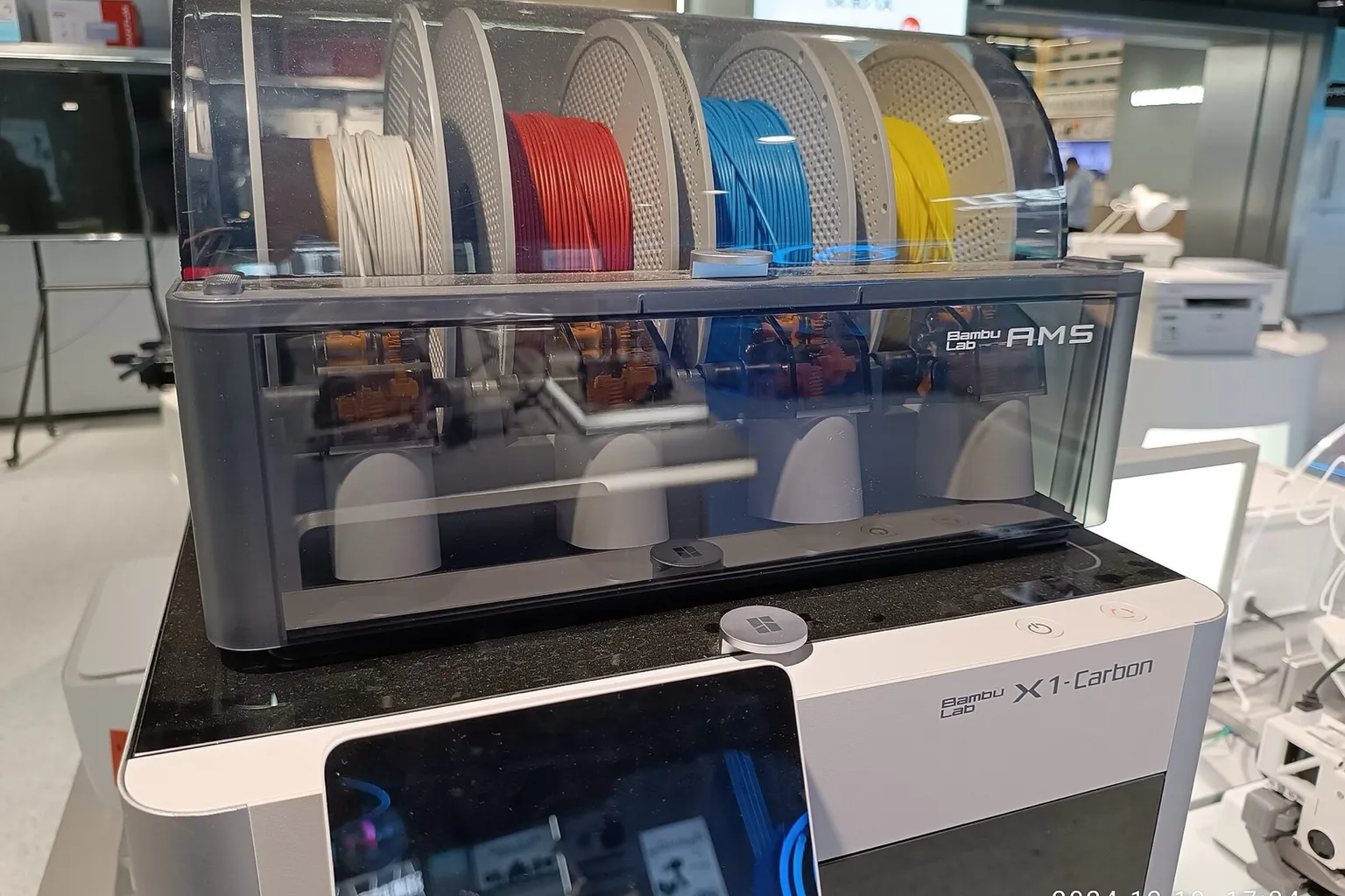 A Bambu Lab AMS holding white, red, blue and yellow filament spools on top of an X1-Carbon printer in a store showroom
