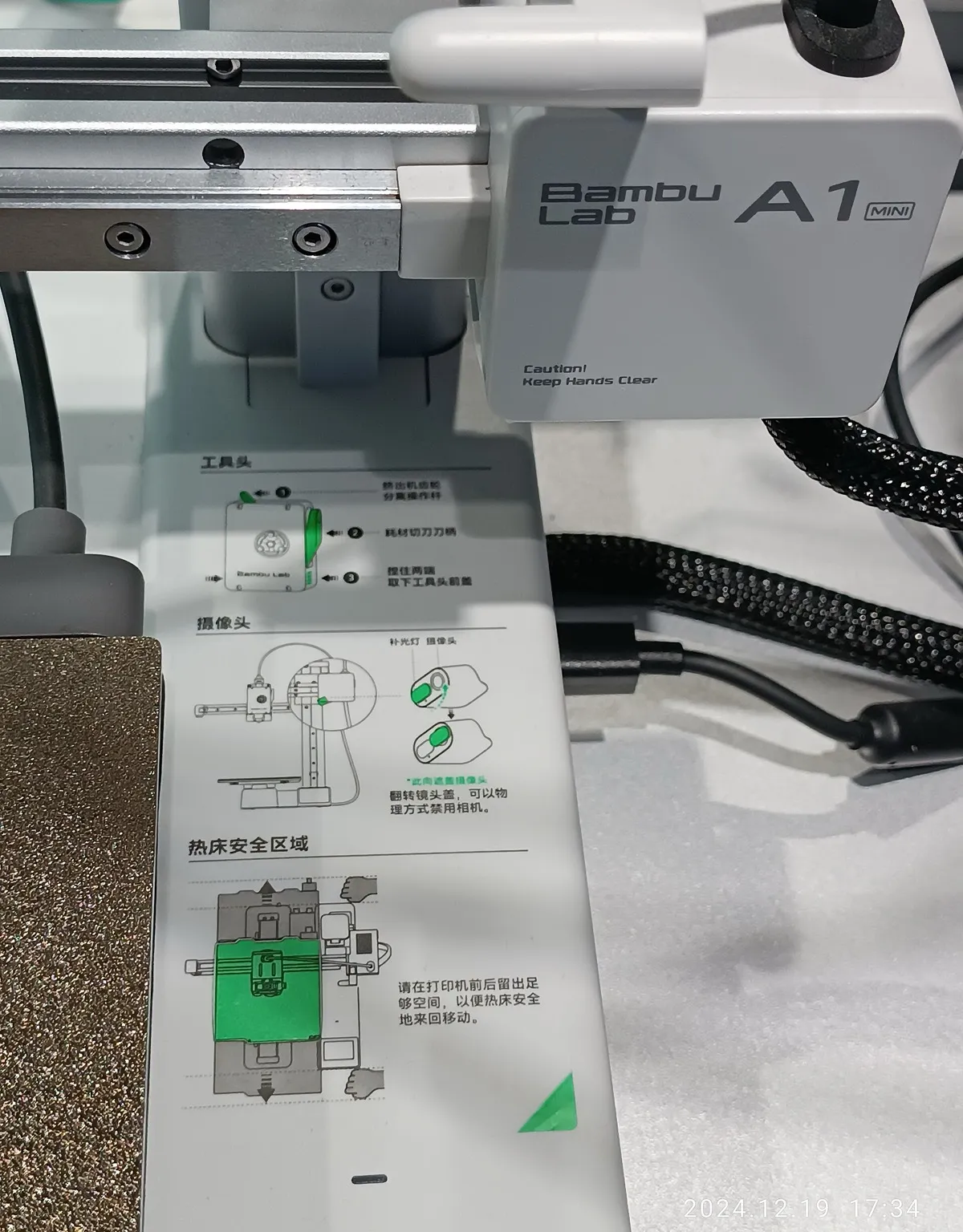 Close-up of a Bambu Lab A1 mini in a store showroom: the toolhead on its gantry, cables, and a Chinese-language label on the base with diagrams of the toolhead, camera and bed safety area