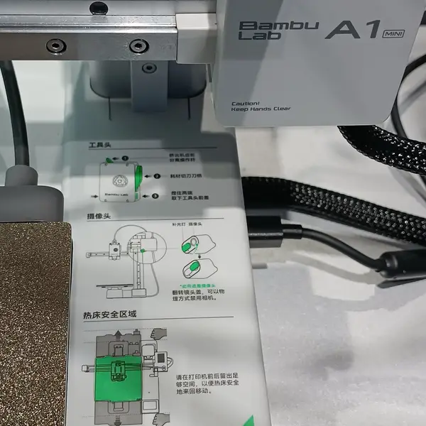 Close-up of a Bambu Lab A1 mini in a store showroom: the toolhead on its gantry, cables, and a Chinese-language label on the base with diagrams of the toolhead, camera and bed safety area