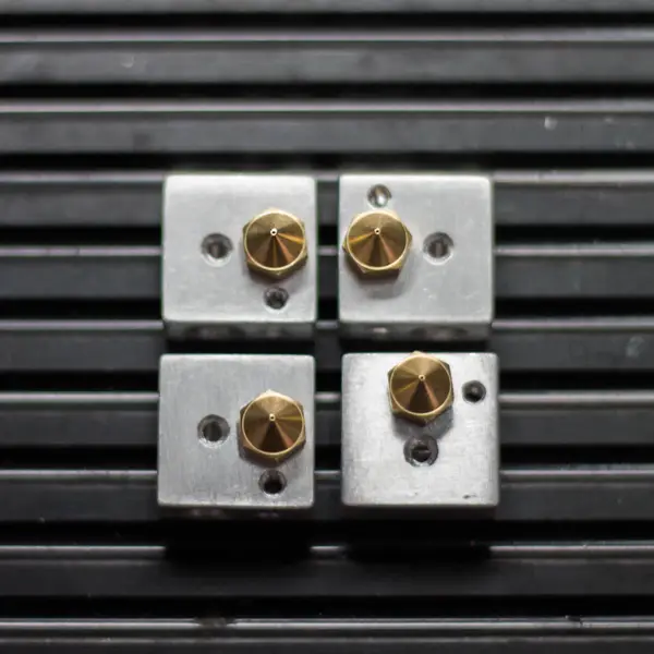 Four aluminum hotend heater blocks in a two-by-two grid on a ribbed black mat, each with a brass nozzle threaded into it, seen from the nozzle end