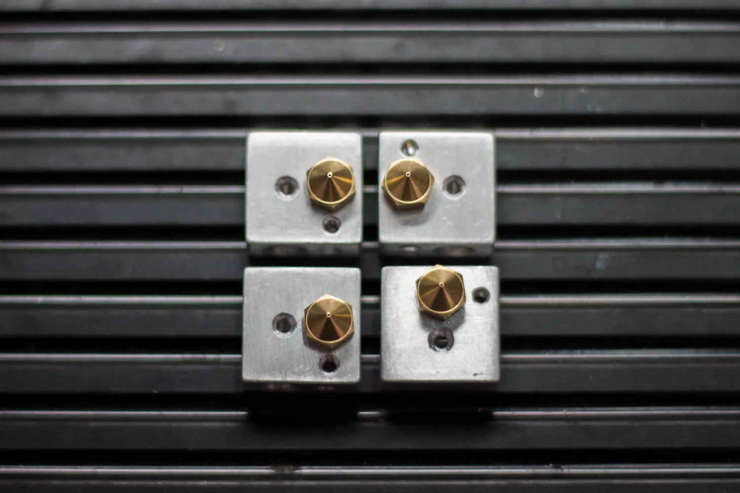 Four aluminum hotend heater blocks in a two-by-two grid on a ribbed black mat, each with a brass nozzle threaded into it, seen from the nozzle end