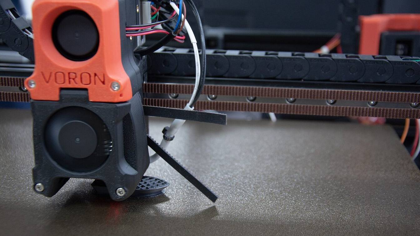 An orange Voron toolhead with its cooling fans, low over a textured build plate