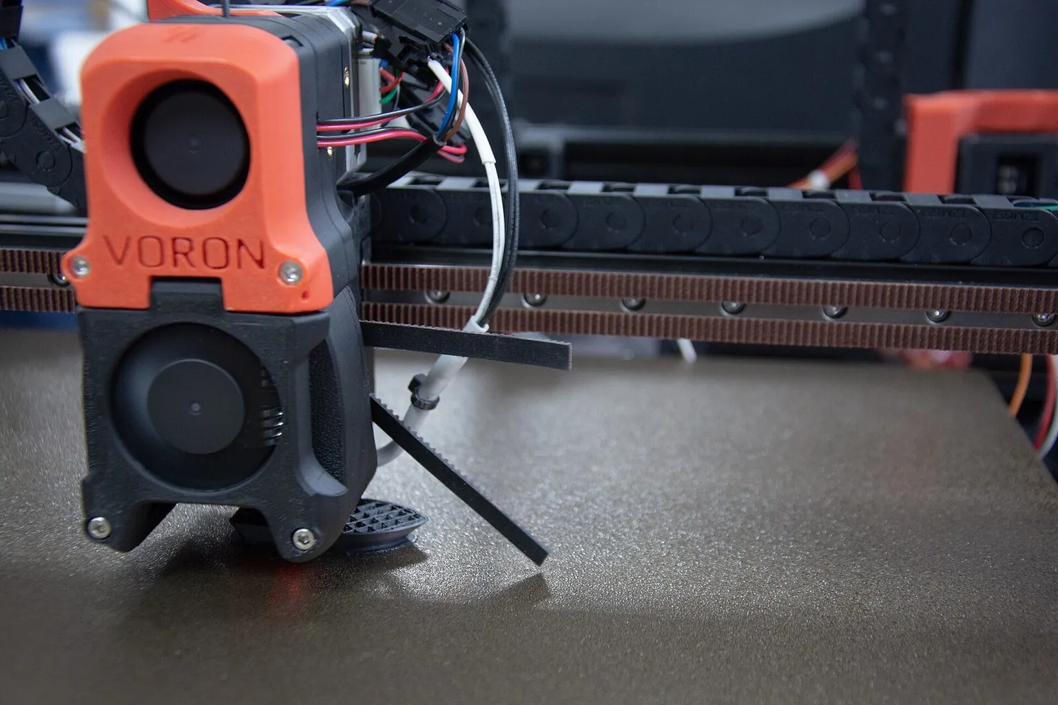 An orange Voron toolhead with its cooling fans, low over a textured build plate