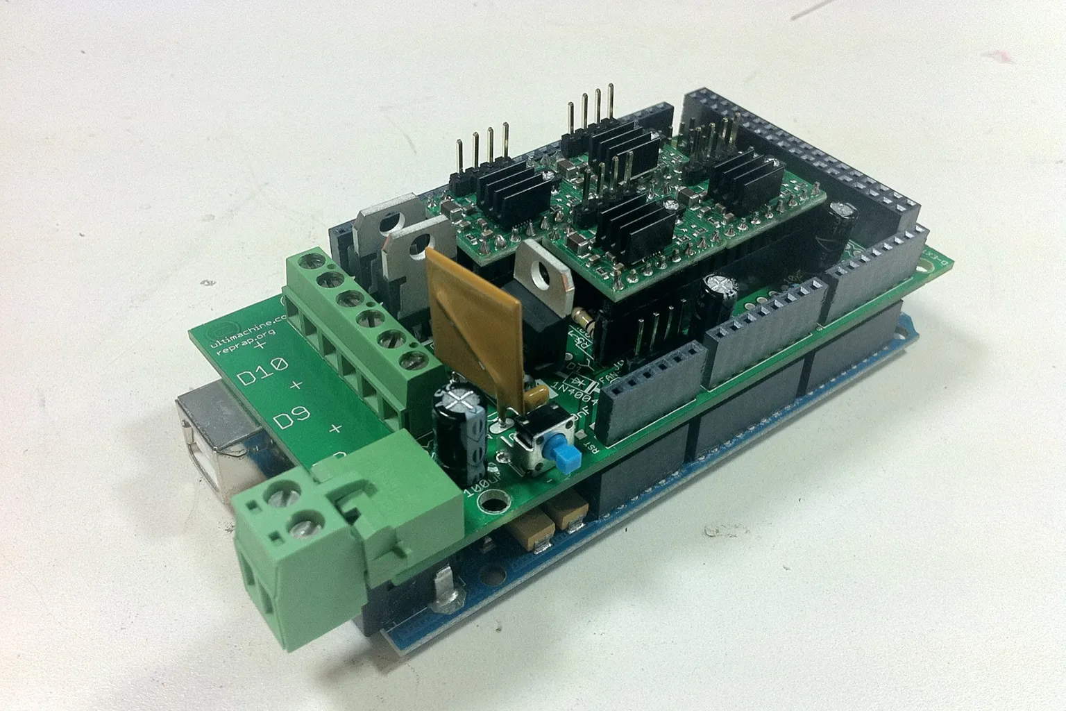 A RAMPS 3D printer controller board stacked on an Arduino Mega, with four stepper driver modules, heatsinks and green screw terminals, on a white desk