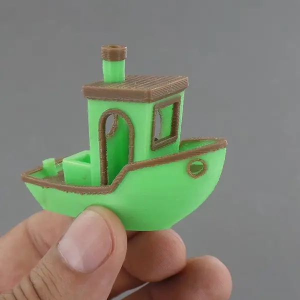 A hand holding a 3DBenchy boat printed in two colors, green and brown
