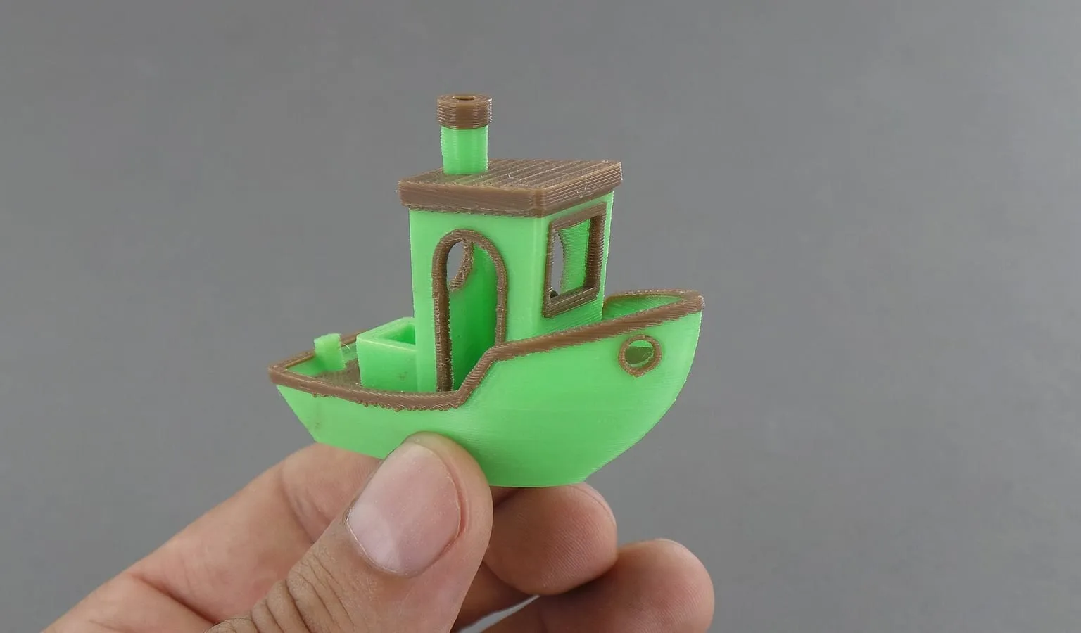 A hand holding a 3DBenchy boat printed in two colors, green and brown