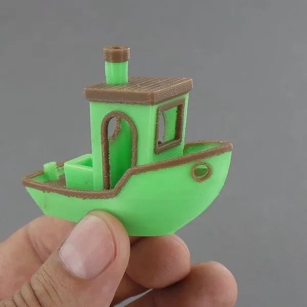 A hand holding a 3DBenchy boat printed in two colors, green and brown