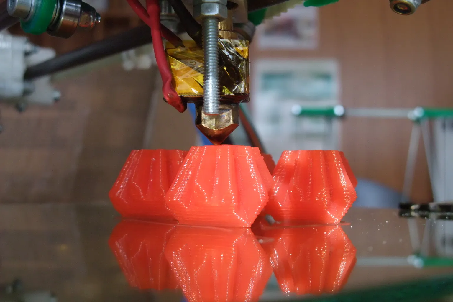 Close-up of a brass nozzle under a Kapton-taped heater block on a RepRap print head, printing three red faceted parts on a glass bed