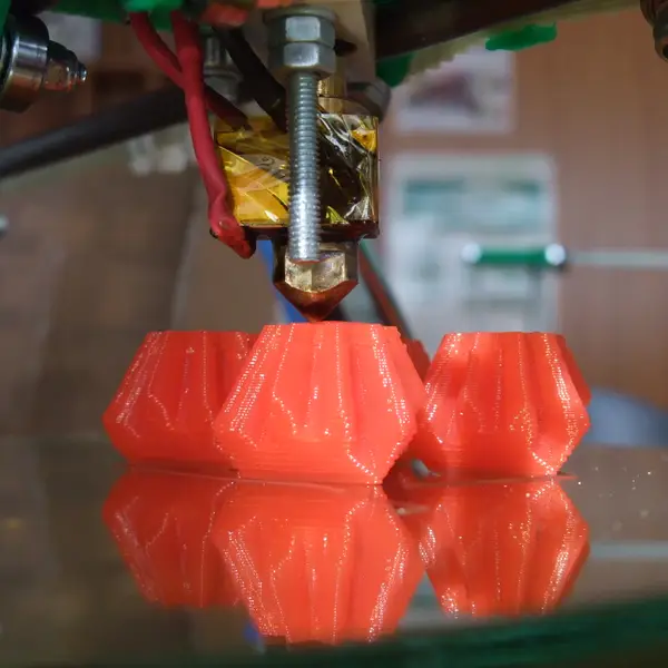 Close-up of a brass nozzle under a Kapton-taped heater block on a RepRap print head, printing three red faceted parts on a glass bed