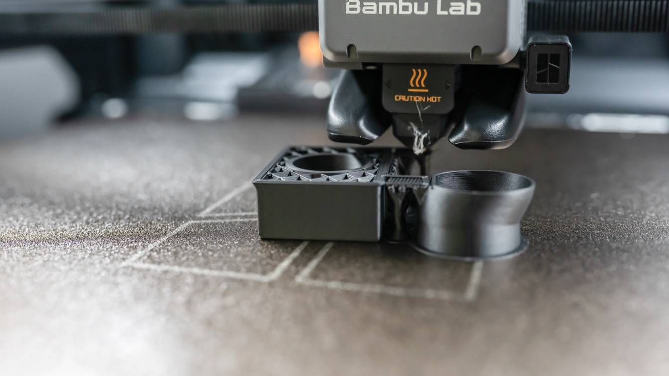 Close-up of a Bambu Lab X1 Carbon toolhead printing two black parts on a textured build plate, the round part ringed by a thin brim at its base