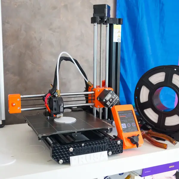 An orange and black Original Prusa MINI on a white shelf printing a small white disc, its orange display showing the job at 47 percent, with a spool of white filament beside it