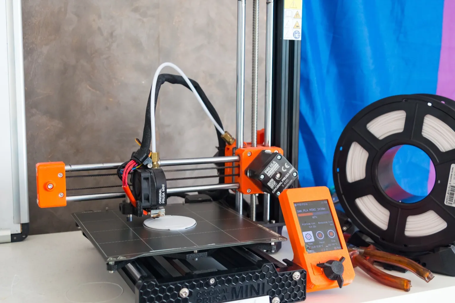 An orange and black Original Prusa MINI on a white shelf printing a small white disc, its orange display showing the job at 47 percent, with a spool of white filament beside it