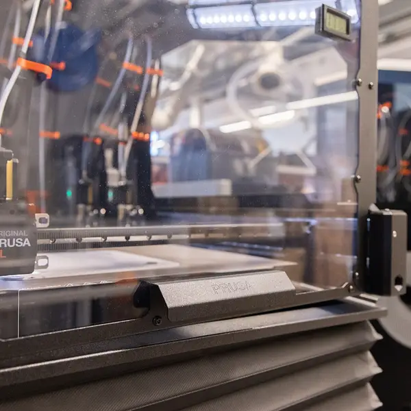 An enclosed Prusa XL in a lab, seen from the side, with the toolhead on the gantry, several PTFE tubes running to the back of the frame and a filament spool on the side