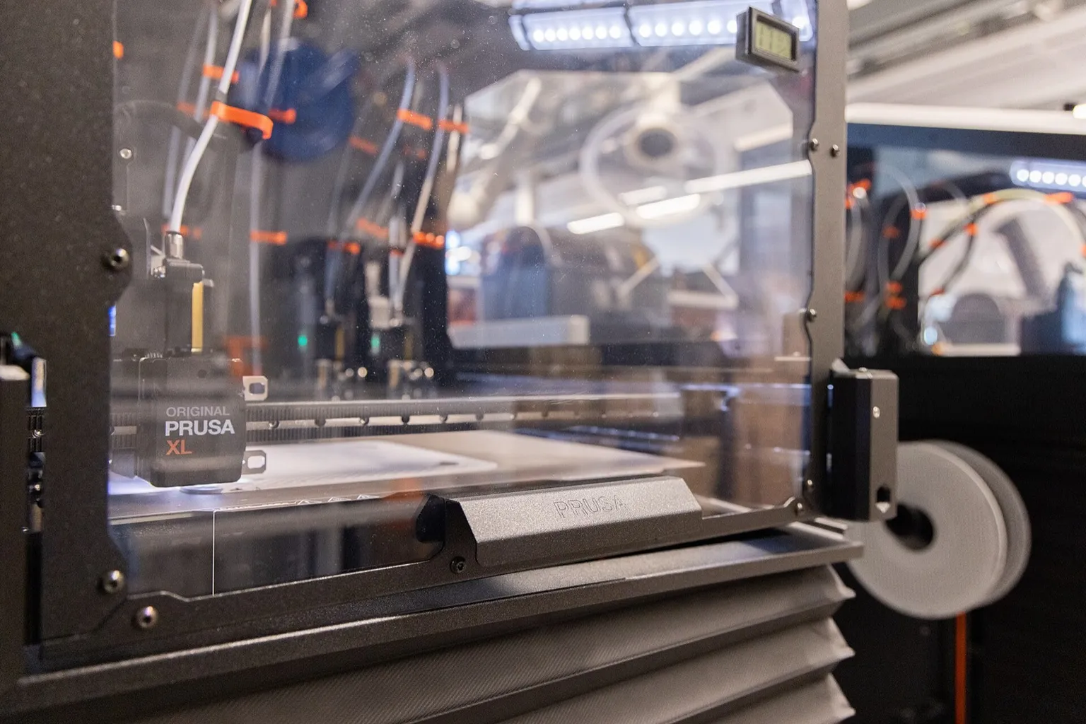 An enclosed Prusa XL in a lab, seen from the side, with the toolhead on the gantry, several PTFE tubes running to the back of the frame and a filament spool on the side