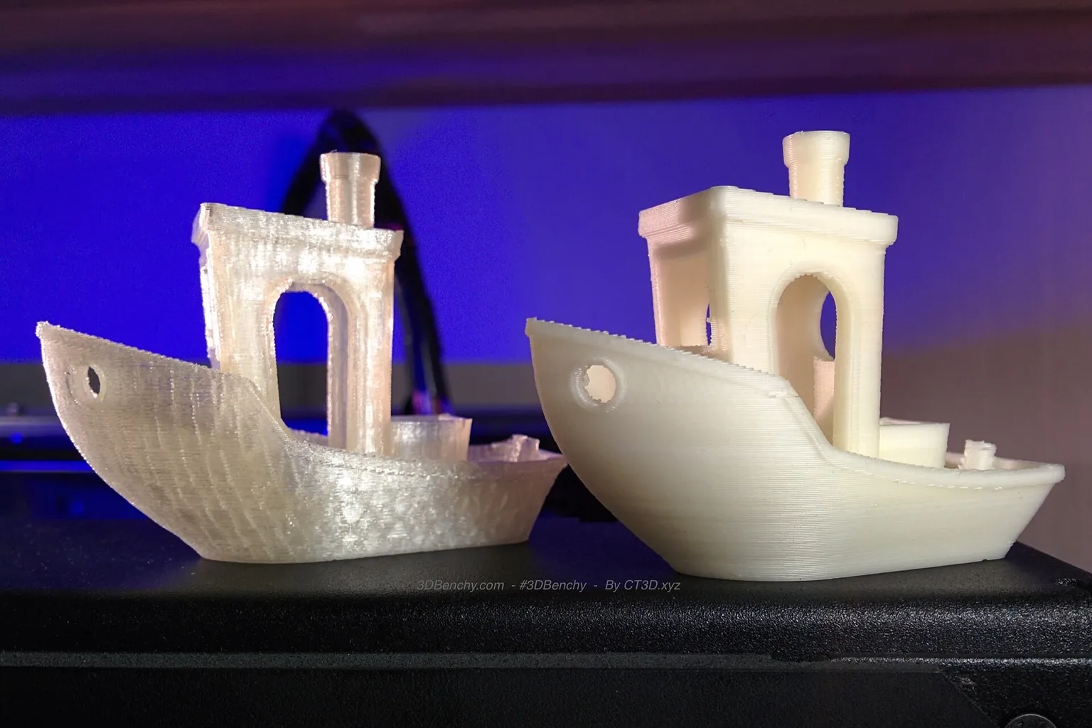 Two 3DBenchy test boats, one translucent and one cream-colored, printed in water-soluble PVA and standing on a black printer top