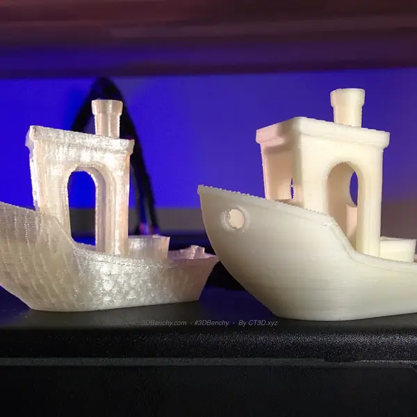 Two 3DBenchy test boats, one translucent and one cream-colored, printed in water-soluble PVA and standing on a black printer top