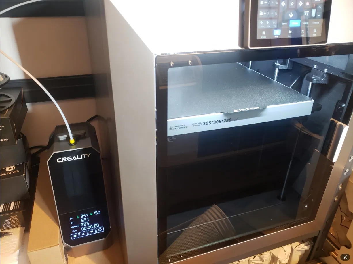 A grey QIDI 3D printer with its door closed and a build plate marked 305 x 305 x 280 inside, next to a Creality filament dryer feeding filament through a white tube