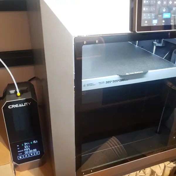 A grey QIDI 3D printer with its door closed and a build plate marked 305 x 305 x 280 inside, next to a Creality filament dryer feeding filament through a white tube