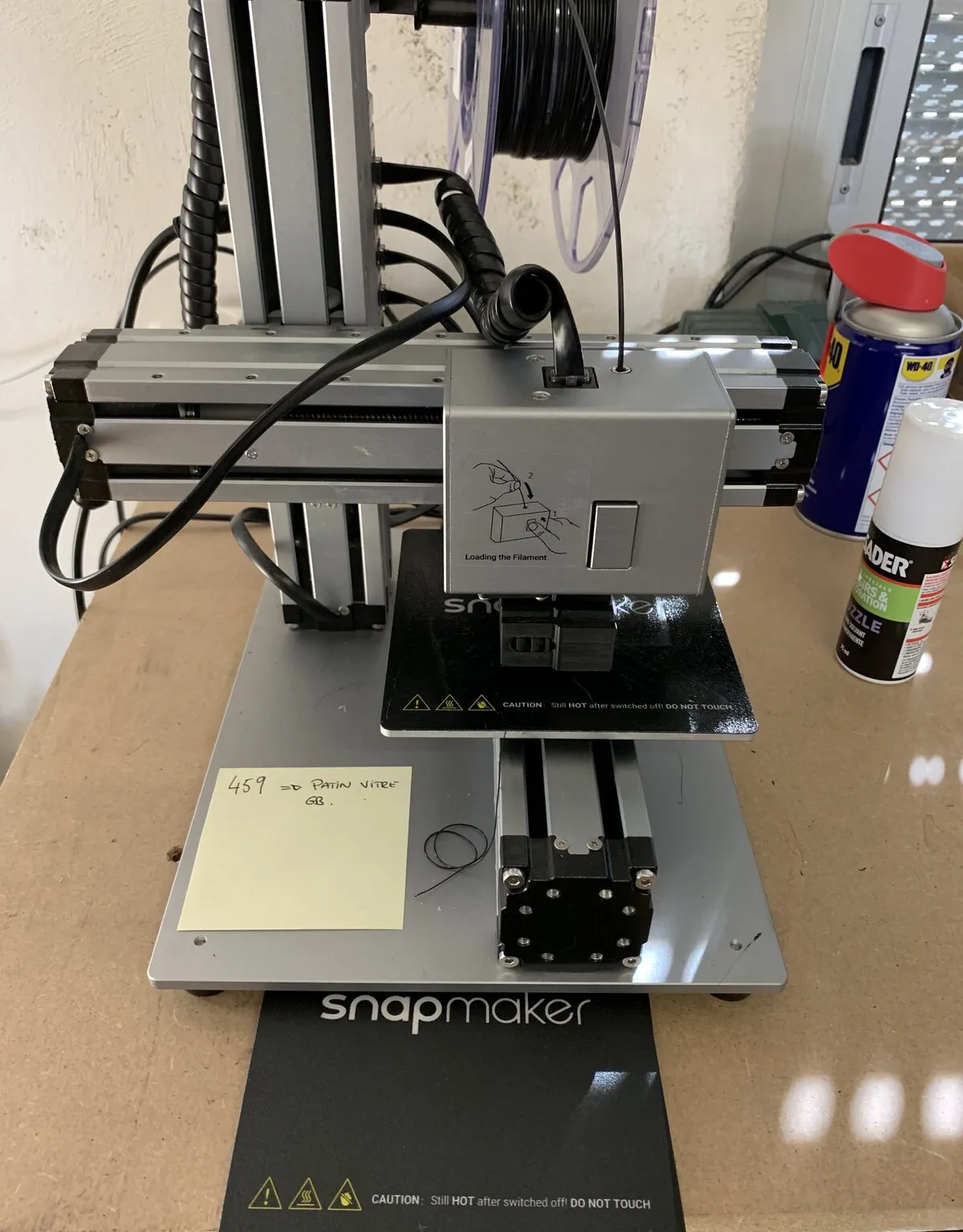 An older single-head Snapmaker 3D printer with an aluminum frame and a filament spool on top, standing on a workbench next to a spray can