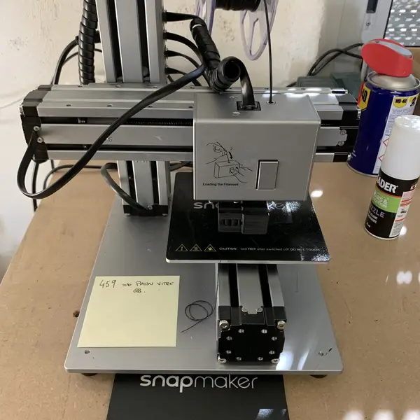 An older single-head Snapmaker 3D printer with an aluminum frame and a filament spool on top, standing on a workbench next to a spray can