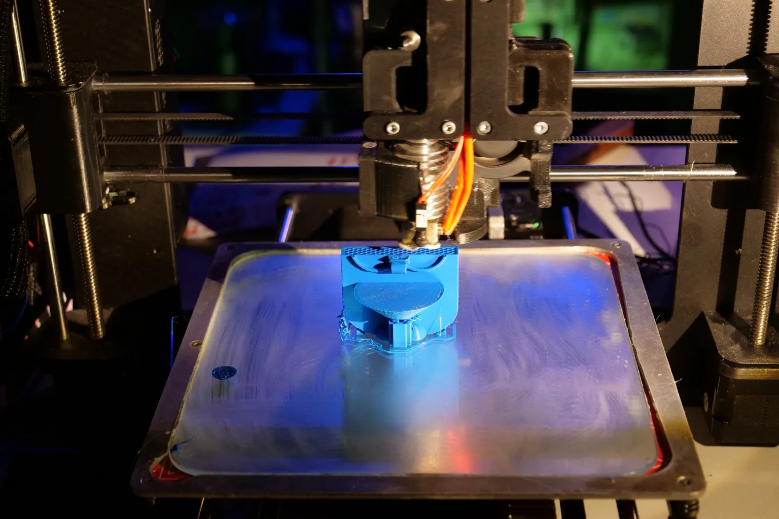 An open-frame i3-style 3D printer, lit in blue, printing a blue part on the glass plate of its heated bed