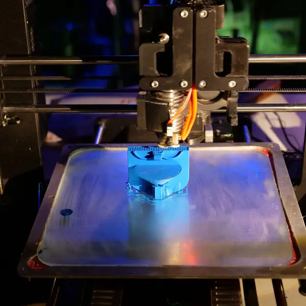 An open-frame i3-style 3D printer, lit in blue, printing a blue part on the glass plate of its heated bed