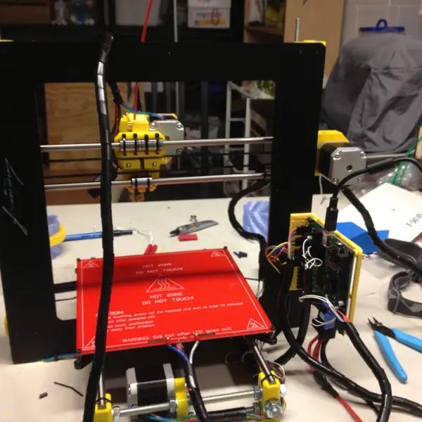 A partly built Prusa i3 printer on a workbench, with its wiring bundled in black spiral wrap running from the electronics board to the print carriage, and wire cutters beside it