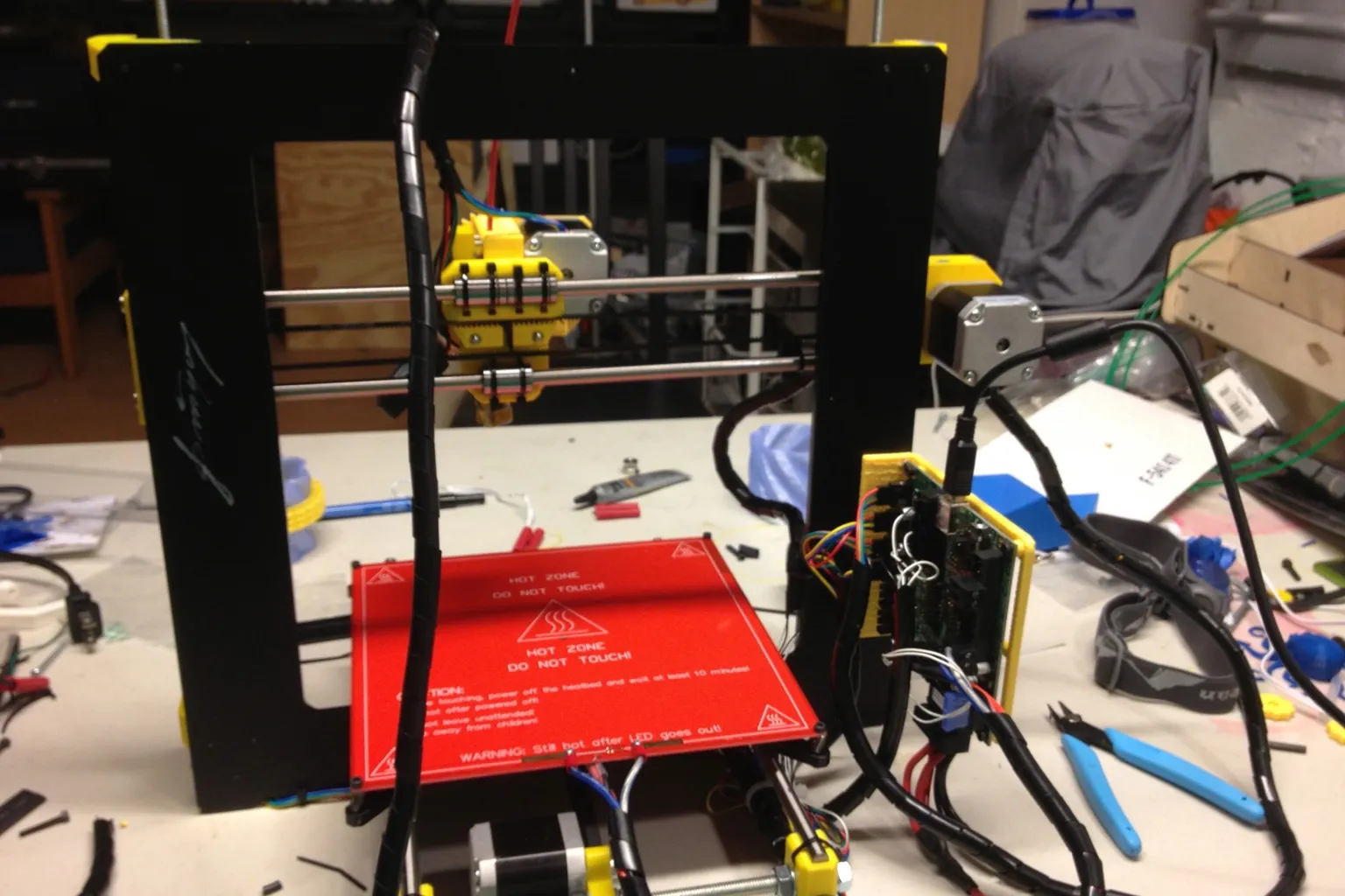 A partly built Prusa i3 printer on a workbench, with its wiring bundled in black spiral wrap running from the electronics board to the print carriage, and wire cutters beside it