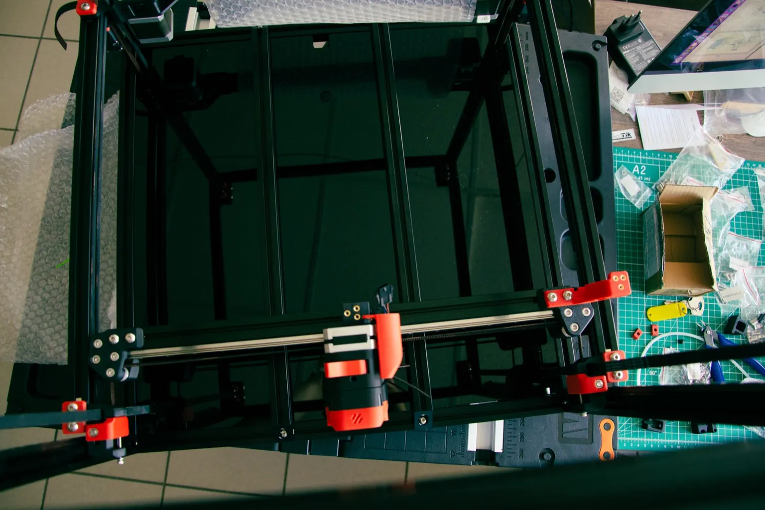 Top-down view of a Voron 2.4 frame under assembly, with a red and black toolhead on the X gantry above the empty bed area