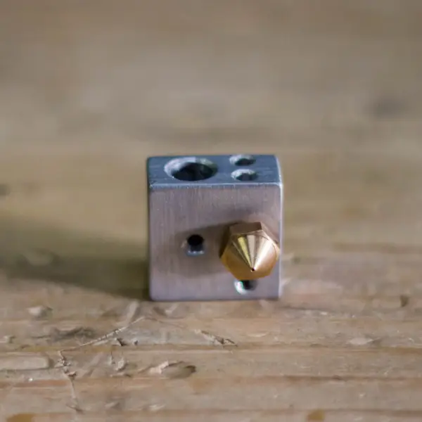 An aluminum hotend heater block with a brass nozzle threaded into it, sitting on a wooden table