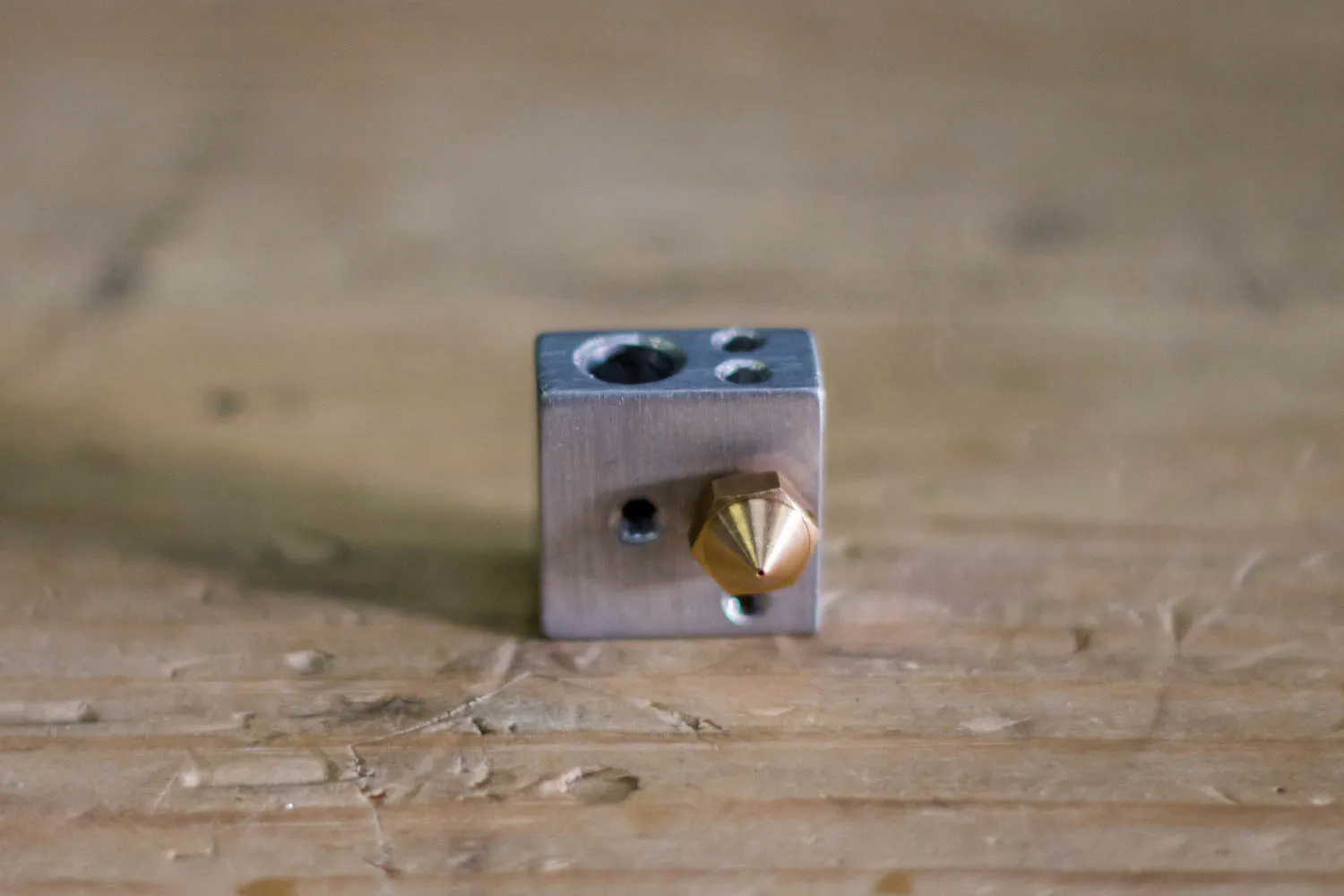 An aluminum hotend heater block with a brass nozzle threaded into it, sitting on a wooden table