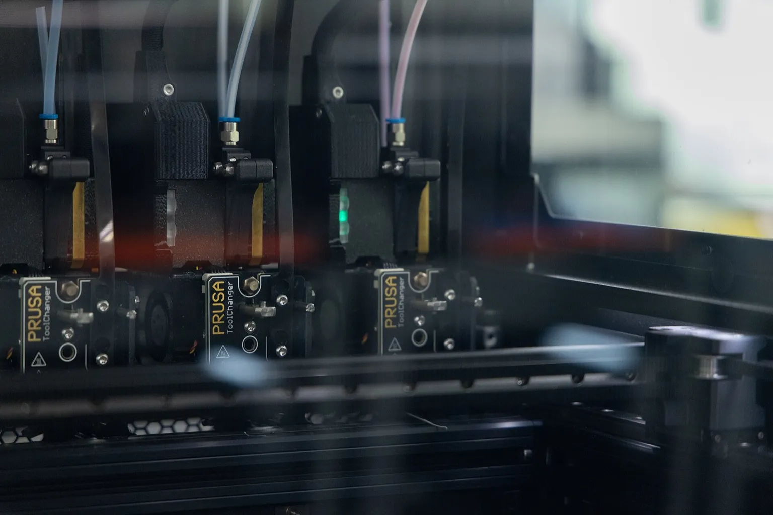 Three Prusa XL toolheads labelled Prusa ToolChanger parked side by side in their docks at the back of the printer, seen through the clear enclosure panel, with filament tubes running up from each one