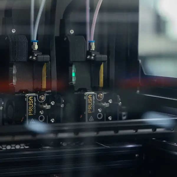 Three Prusa XL toolheads labelled Prusa ToolChanger parked side by side in their docks at the back of the printer, seen through the clear enclosure panel, with filament tubes running up from each one