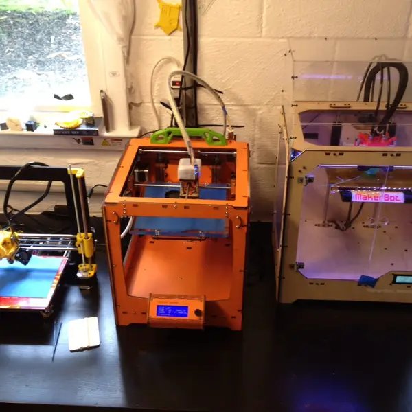 Three hobbyist 3D printers side by side on a desk against a white brick wall: an open-frame Prusa i3, an orange plywood-frame printer and a MakerBot in a wooden case with clear panels