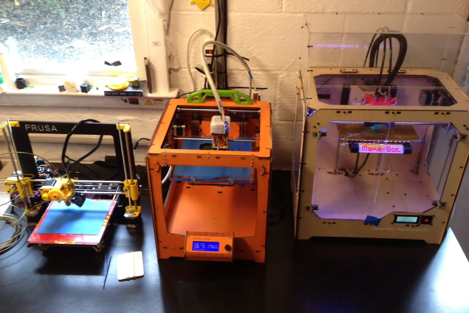 Three hobbyist 3D printers side by side on a desk against a white brick wall: an open-frame Prusa i3, an orange plywood-frame printer and a MakerBot in a wooden case with clear panels