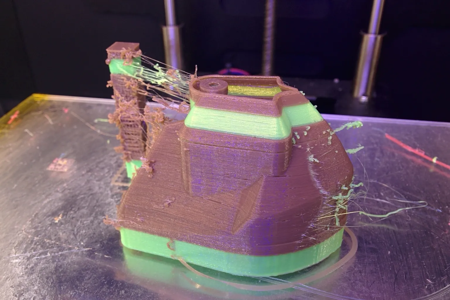 A brown and green two-color 3D-printed boat on a glass bed, with a strung-up purge tower beside it and thin green strings of oozed filament across the print