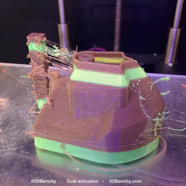A brown and green two-color 3D-printed boat on a glass bed, with a strung-up purge tower beside it and thin green strings of oozed filament across the print