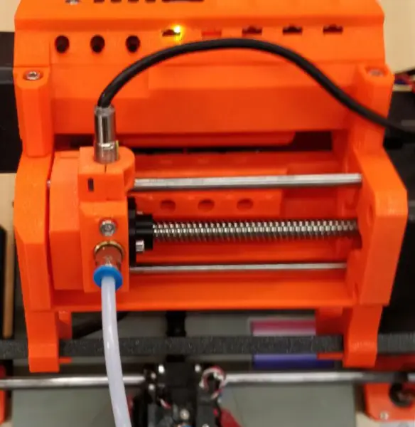Orange Prusa Multi-Material Upgrade unit mounted on top of a Prusa i3 printer frame: a selector carriage on a lead screw and steel rods, with a white PTFE tube running down to the extruder