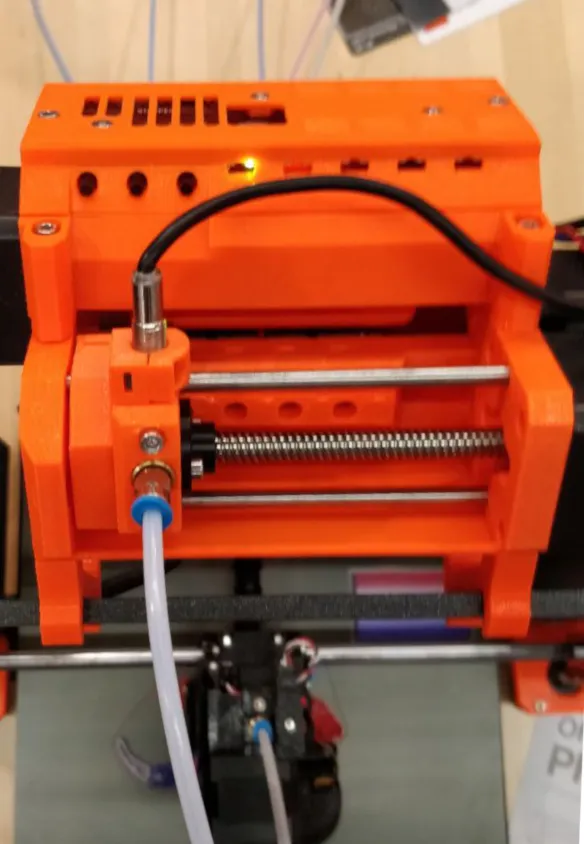 Orange Prusa Multi-Material Upgrade unit mounted on top of a Prusa i3 printer frame: a selector carriage on a lead screw and steel rods, with a white PTFE tube running down to the extruder