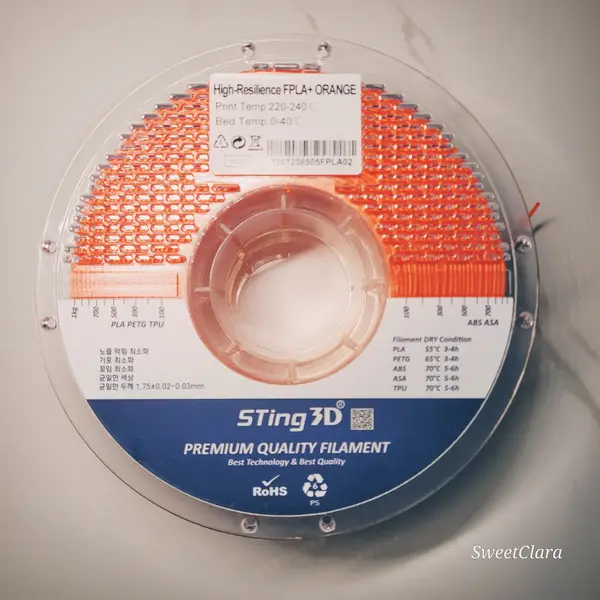 A clear plastic spool of orange flexible filament with a label listing print and bed temperatures, on a marble surface