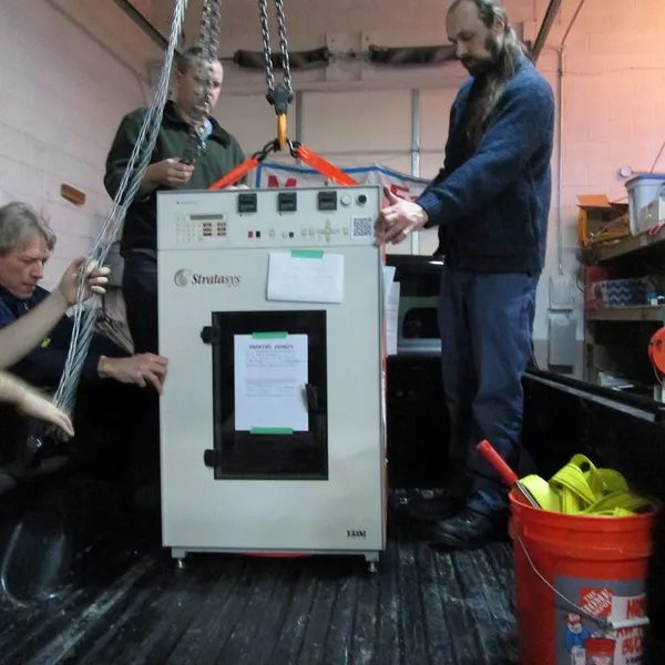 Three people hoisting a cabinet-sized Stratasys FDM machine off a truck