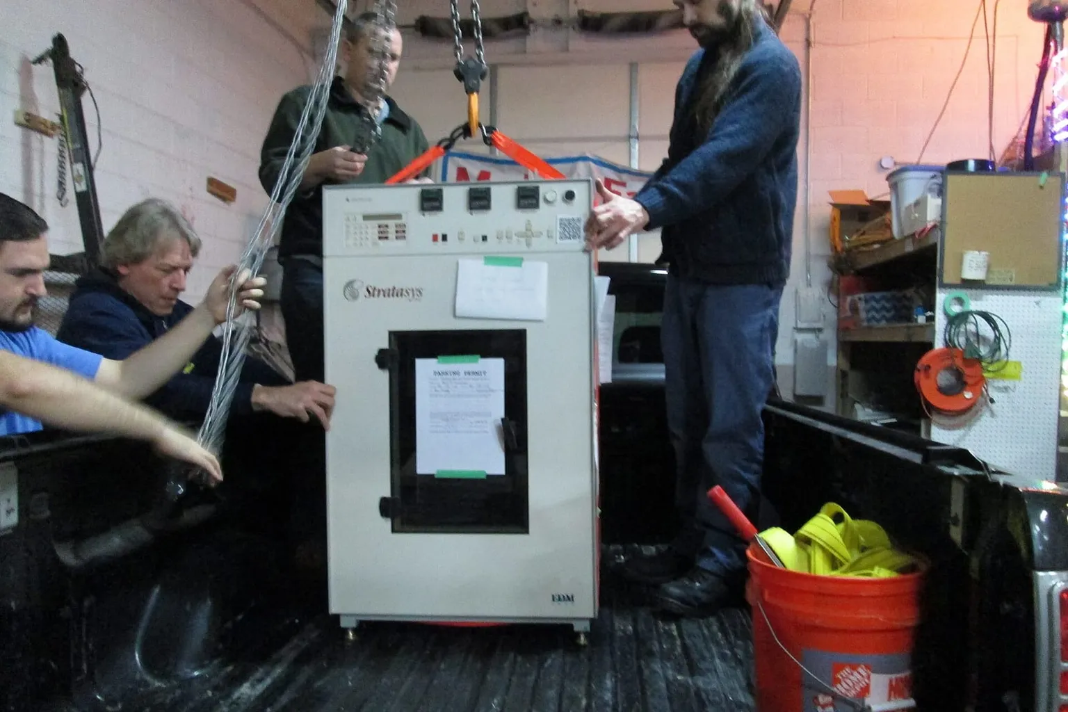 Three people hoisting a cabinet-sized Stratasys FDM machine off a truck
