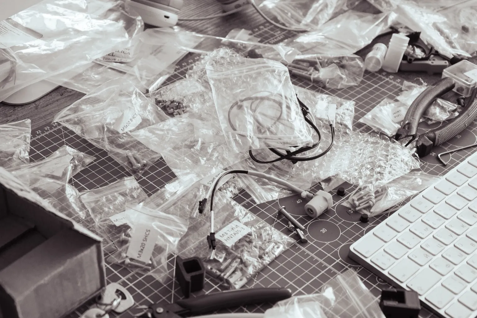 Black-and-white photo of Voron printer kit parts on a cutting mat: labelled bags of M3 and M5 screws, wiring, printed parts, pliers and a keyboard
