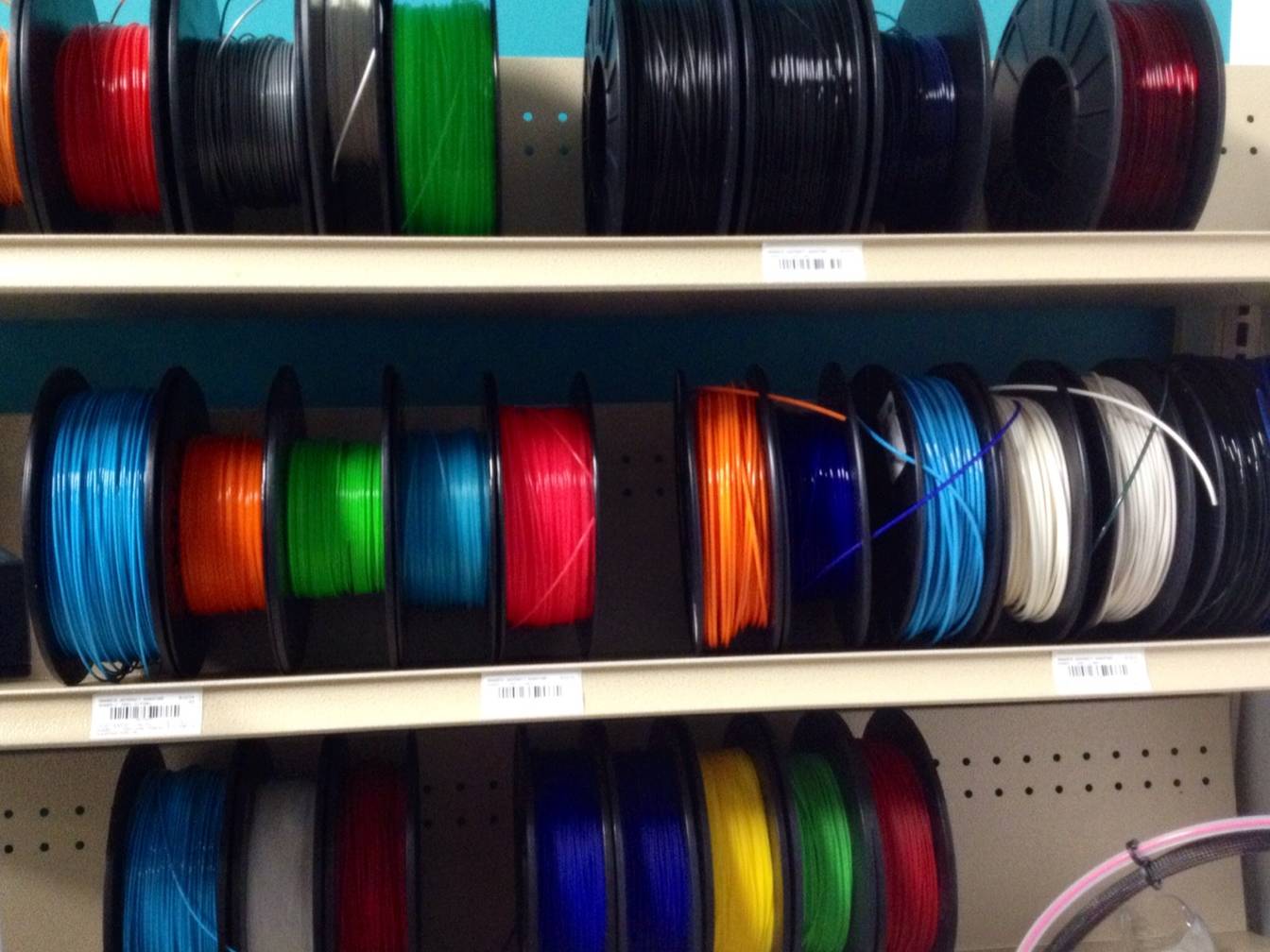 Three shelves of 3D printer filament spools in many colors, among them red, black, green, blue, orange, white and yellow, stored side by side in a maker lab